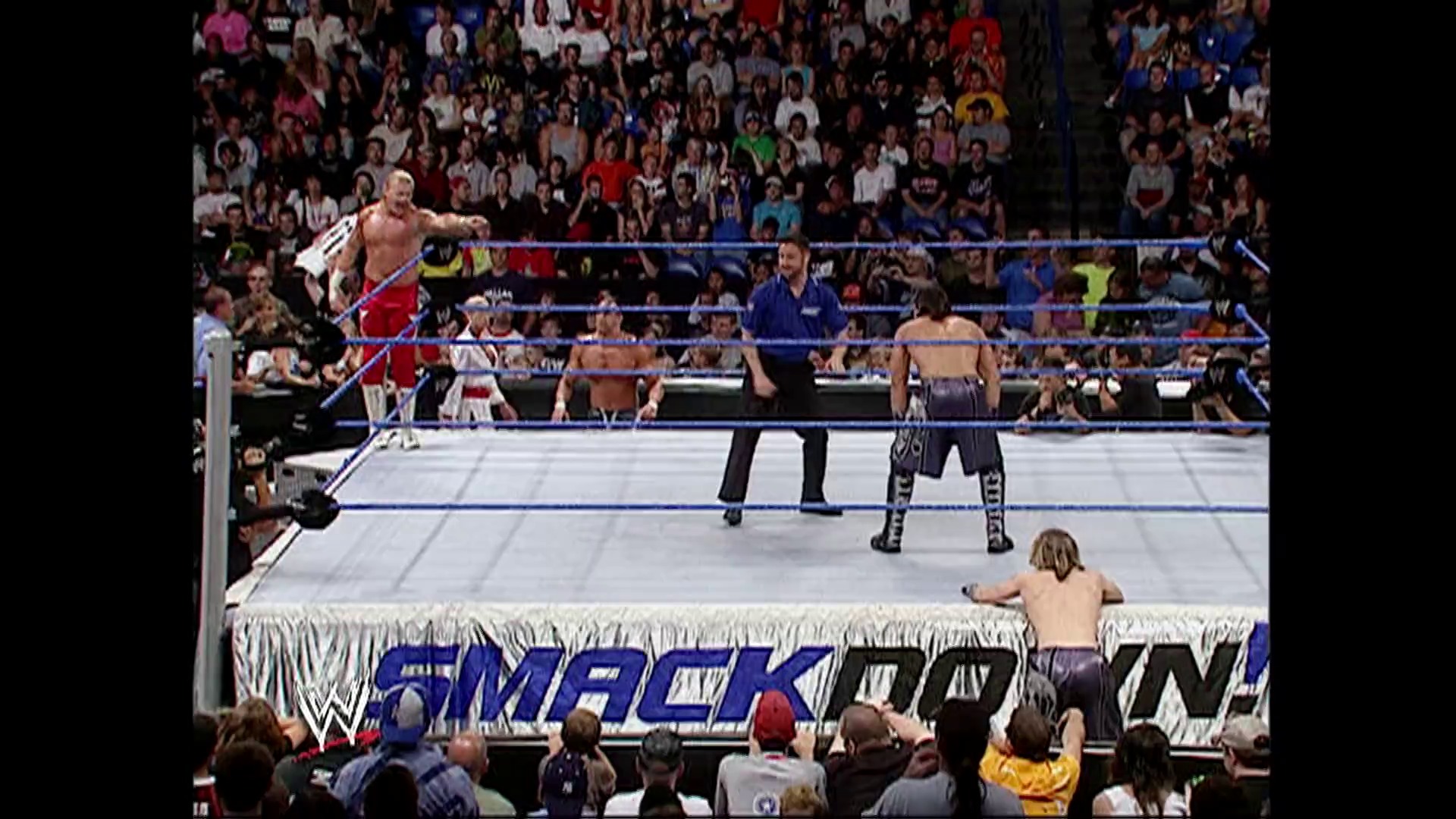 A frame from WWE SmackDown (August 25, 2006) at 51:18. A WWE SmackDown wrestling match in progress with multiple wrestlers in the ring, a referee, and a packed crowd visible in the background. The blue-roped ring displays the SmackDown branding, and one wrestler appears to be performing an aerial move.