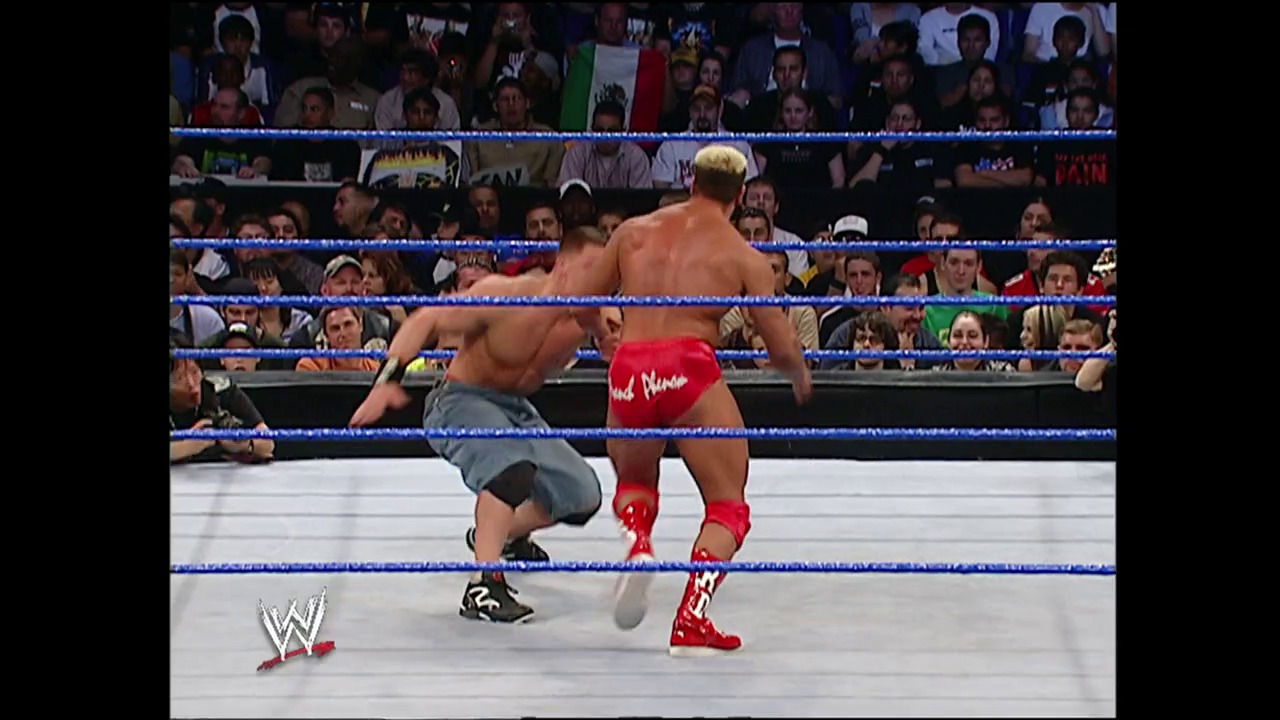 A frame from WWE Judgment Day (2004) at 1:29:26. A WWE wrestler in red and white attire faces off against an opponent in gray shorts in the blue-roped ring, while a packed crowd watches from the stands. The WWE logo is visible in the lower left corner of the broadcast.