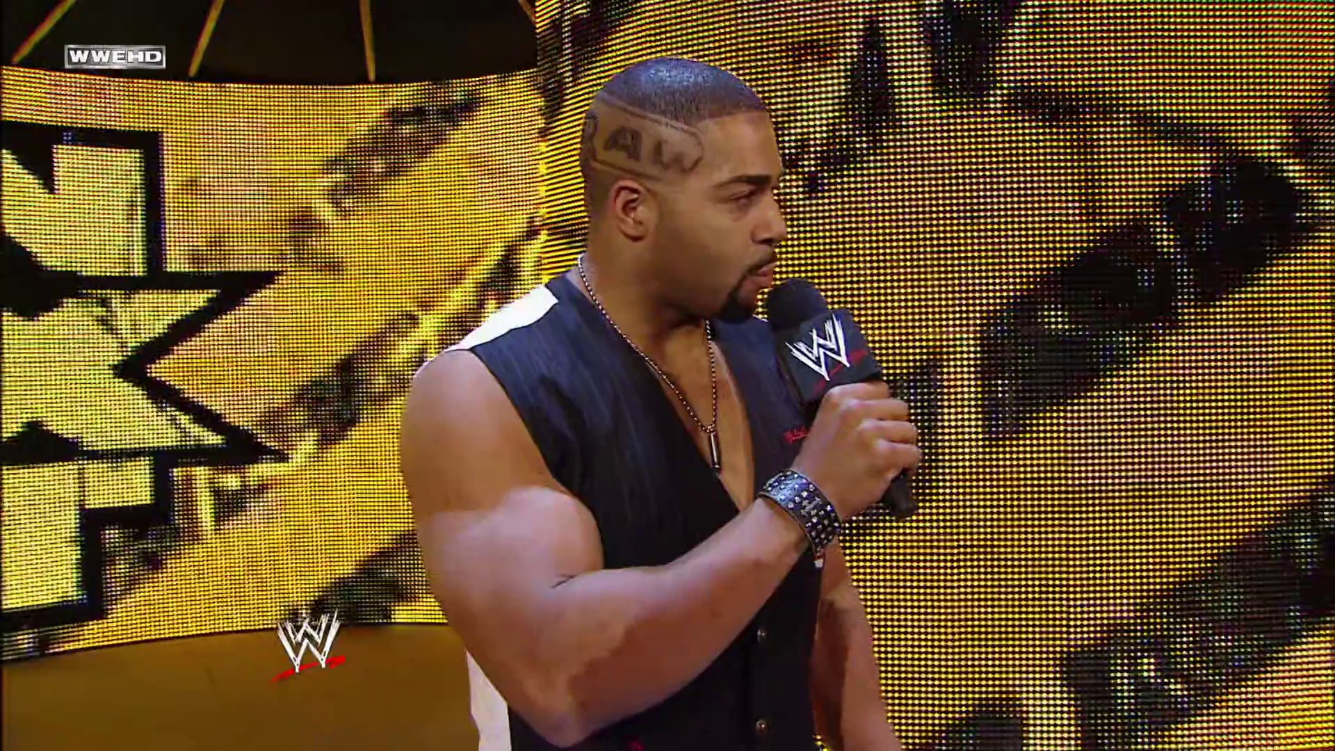 A frame from WWE Raw (April 5, 2010) at 13:28. A WWE wrestler stands in the ring holding a microphone, wearing a dark sleeveless vest and chain necklace. Behind him, a large yellow LED screen displays WWE branding while the WWE logo appears in the lower corner of the broadcast.