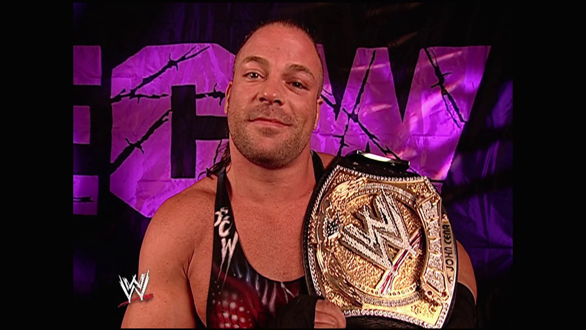 A frame from WWE Raw (June 12, 2006) at 2:54. A professional wrestler stands in the ring wearing a black and red sleeveless shirt, displaying a championship belt across his shoulder. Behind him is a purple-tinted WWE Raw graphic backdrop with decorative branch designs.