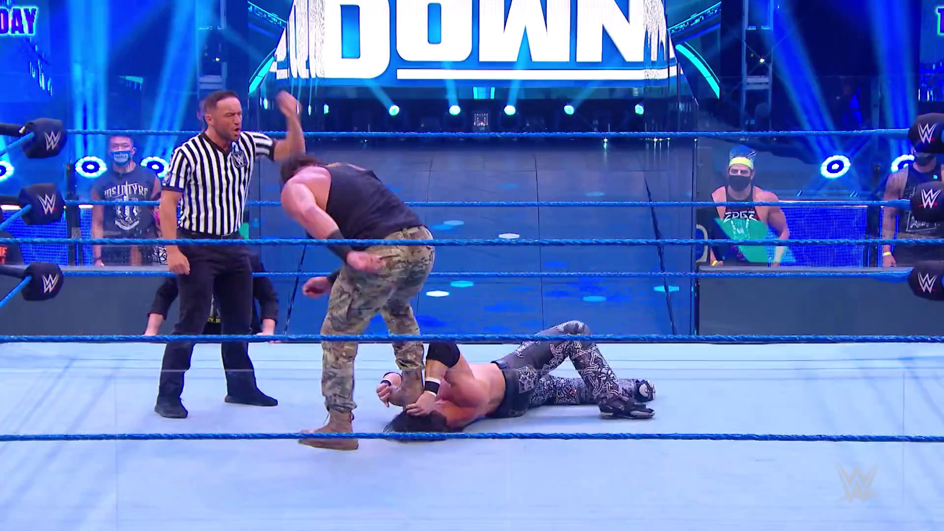 A frame from WWE SmackDown (July 17, 2020) at 47:08. A WWE SmackDown match in progress featuring a wrestler in camouflage pants standing over a fallen opponent on the blue ring, with a referee in striped shirt observing and the illuminated "SMACKDOWN" graphic visible in the background.