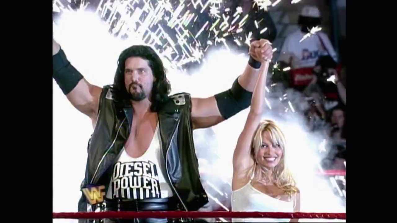 A frame from WrestleMania XI (1995) at 1:37:02. # WrestleMania XI Scene

A long-haired wrestler in a leather vest and "Diesel Power" shirt celebrates with his fist raised, accompanied by a blonde woman, as pyrotechnics explode behind them in a dramatic WWE arena entrance.