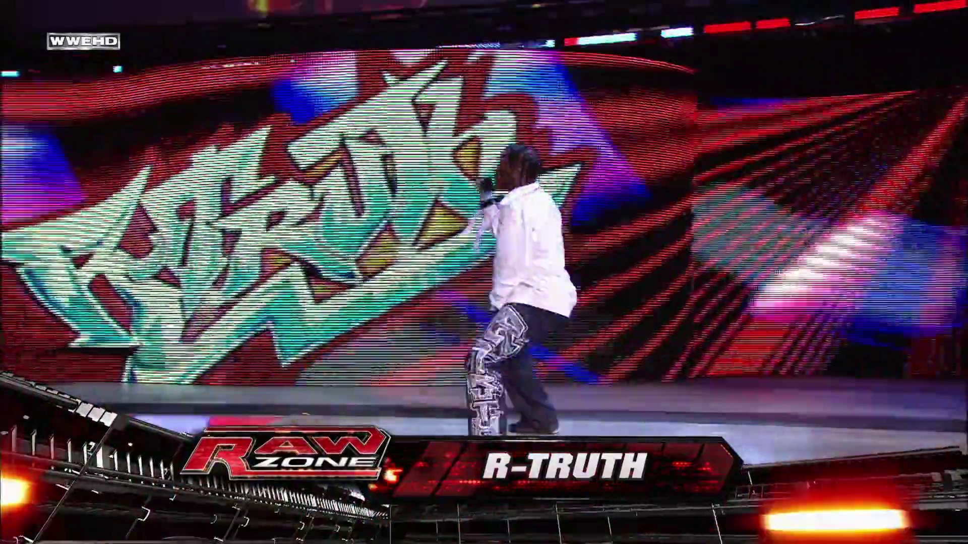 A frame from WWE Raw (November 1, 2010) at 1:15:58. A WWE Raw performer identified as R-Truth stands on the ring apron in a white shirt and patterned black pants, facing a large LED screen displaying colorful graphics with red, blue, and cyan lighting effects typical of WWE's production setup.