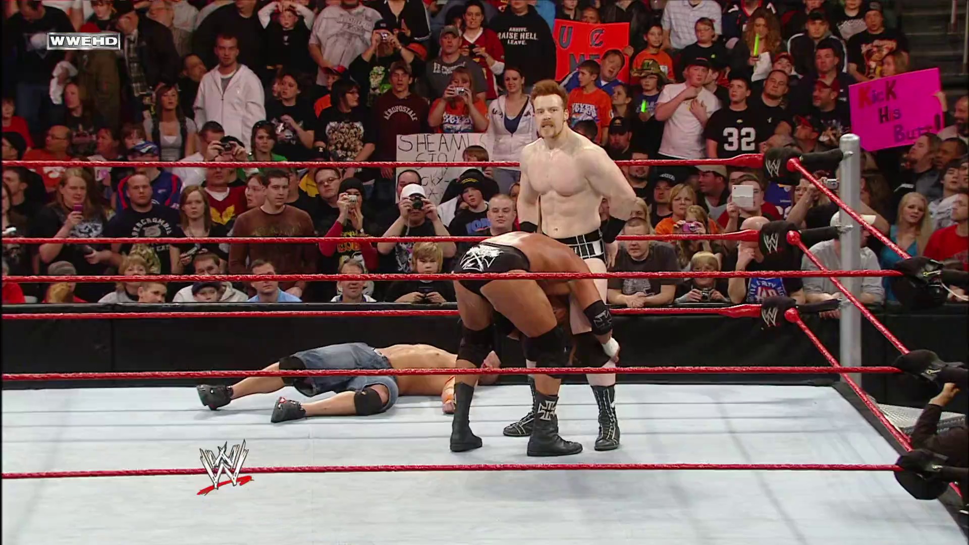 A frame from WWE Raw (February 15, 2010) at 1:29:37. A WWE wrestler stands in the red-roped ring over a downed opponent during Raw. The packed crowd fills the arena behind the ring, with visible signage and the WWE logo displayed on the mat.