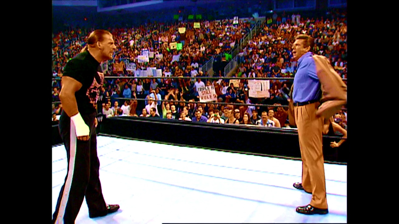 A frame from WWE Raw (May 16, 2002) at 0:54. Two wrestlers face off in a WWE ring before a packed arena crowd. One wears black attire with red accents, while the other sports a blue shirt and tan pants, with spectators holding signs visible in the background.