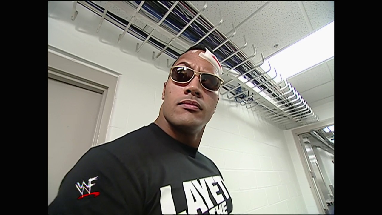 A frame from WWE SmackDown (April 13, 2000) at 1:16:30. A wrestler wearing sunglasses and a black WWF "Layeth the Smacketh Down" t-shirt stands in an industrial-style backstage area with exposed ceiling infrastructure and white walls, captured in a low-angle shot typical of early 2000s WWE production.