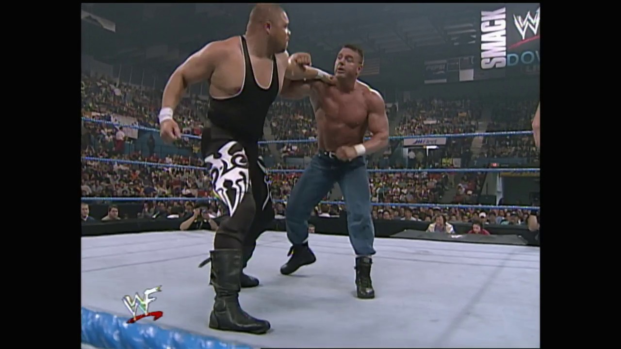 A frame from WWE SmackDown (October 28, 1999) at 37:57. Two wrestlers face off in a WWE SmackDown ring, with one in black attire with tribal designs and the other shirtless in jeans. A packed crowd fills the arena in the background, with WWE graphics and signage visible overhead.