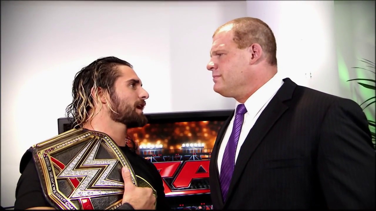 A frame from WWE Raw (April 20, 2015) at 0:56. Two wrestlers face off in conversation, with one holding an ornate gold and red championship belt. The man on the right wears a black suit and purple tie, while the other sports long wet hair and black attire. A wrestling ring and WWE graphics are visible in the background.