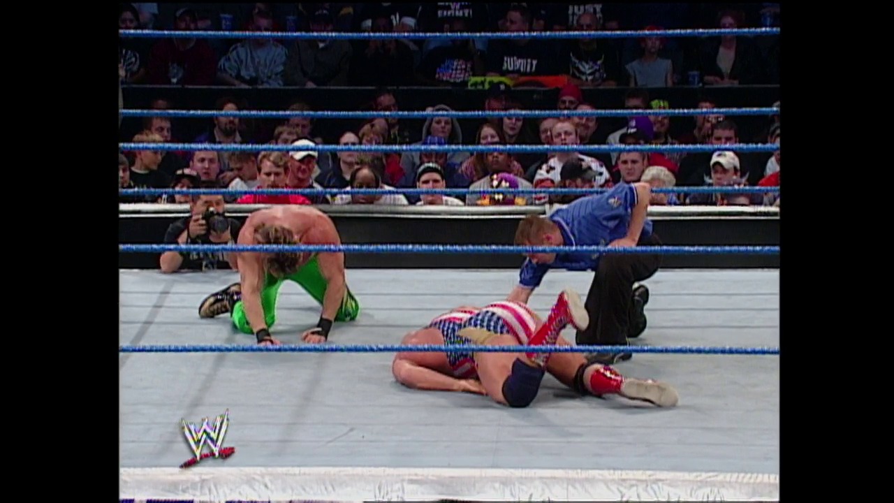 A frame from WWE Raw (October 31, 2002) at 59:20. A WWE wrestler in red, white, and blue attire lies on the mat while another competitor in green stands nearby. The crowd fills the bleachers behind the blue-roped ring, with the WWE logo visible in the corner.