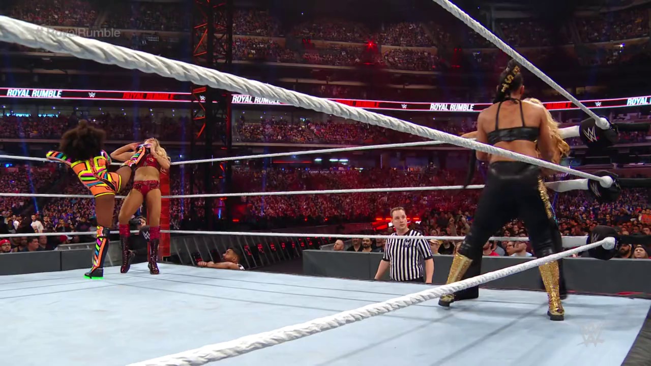 A frame from Royal Rumble (2020) at 1:10:12. Two female wrestlers face off in a WWE ring during Royal Rumble 2020, with one in red and gold attire on the left and another in black and gold on the right, while a referee watches and a packed arena crowd cheers in the background.