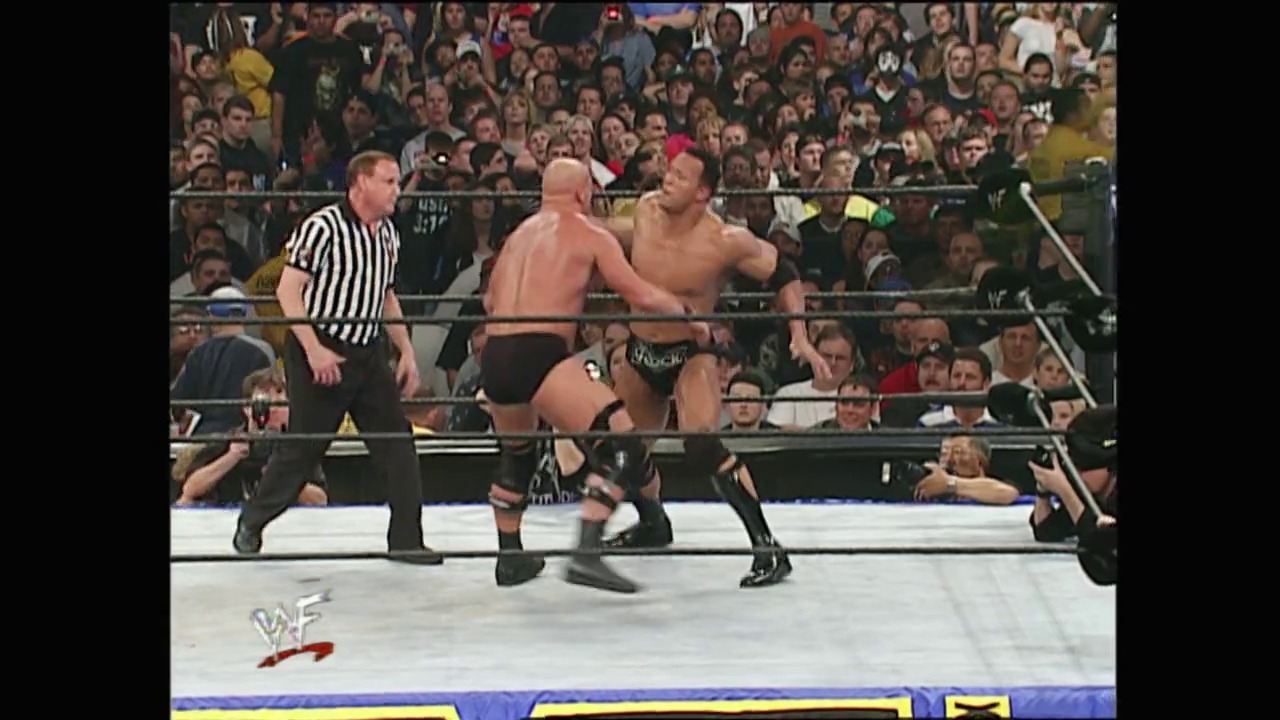 A frame from WrestleMania X-Seven (2001) at 3:13:08. Two shirtless wrestlers face off in a WWF ring while a referee observes, with a packed crowd visible in the background. The white mat, steel ropes, and WWF logo are visible, capturing the intensity of a live wrestling match.