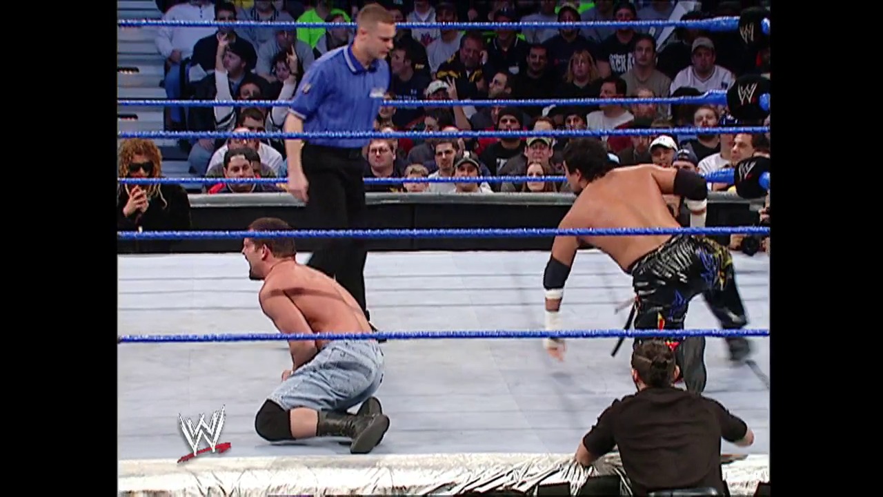A frame from WWE SmackDown (January 15, 2004) at 12:07. A WWE SmackDown match in progress shows a shirtless wrestler on his knees in the ring while opponents and a referee are positioned around him. The packed crowd watches from the stands behind the blue ropes, with the WWE logo visible in the corner.