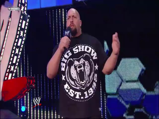 A frame from WWE SmackDown (July 2, 2010) at 12:27. A bald wrestler in a black "Big Show" t-shirt holds a microphone during a WWE SmackDown event, standing on stage with blue LED lighting and graphics visible in the background.