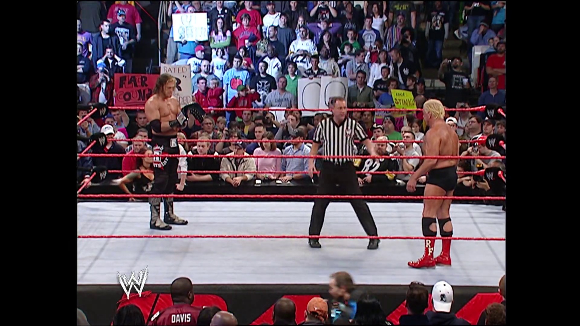 A frame from WWE Raw (January 16, 2006) at 1:15:14. A WWE Raw match in progress features three wrestlers in the red-roped ring with a referee in striped shirt, while a packed arena crowd watches from the stands. The WWE logo is visible at the bottom left of the frame.