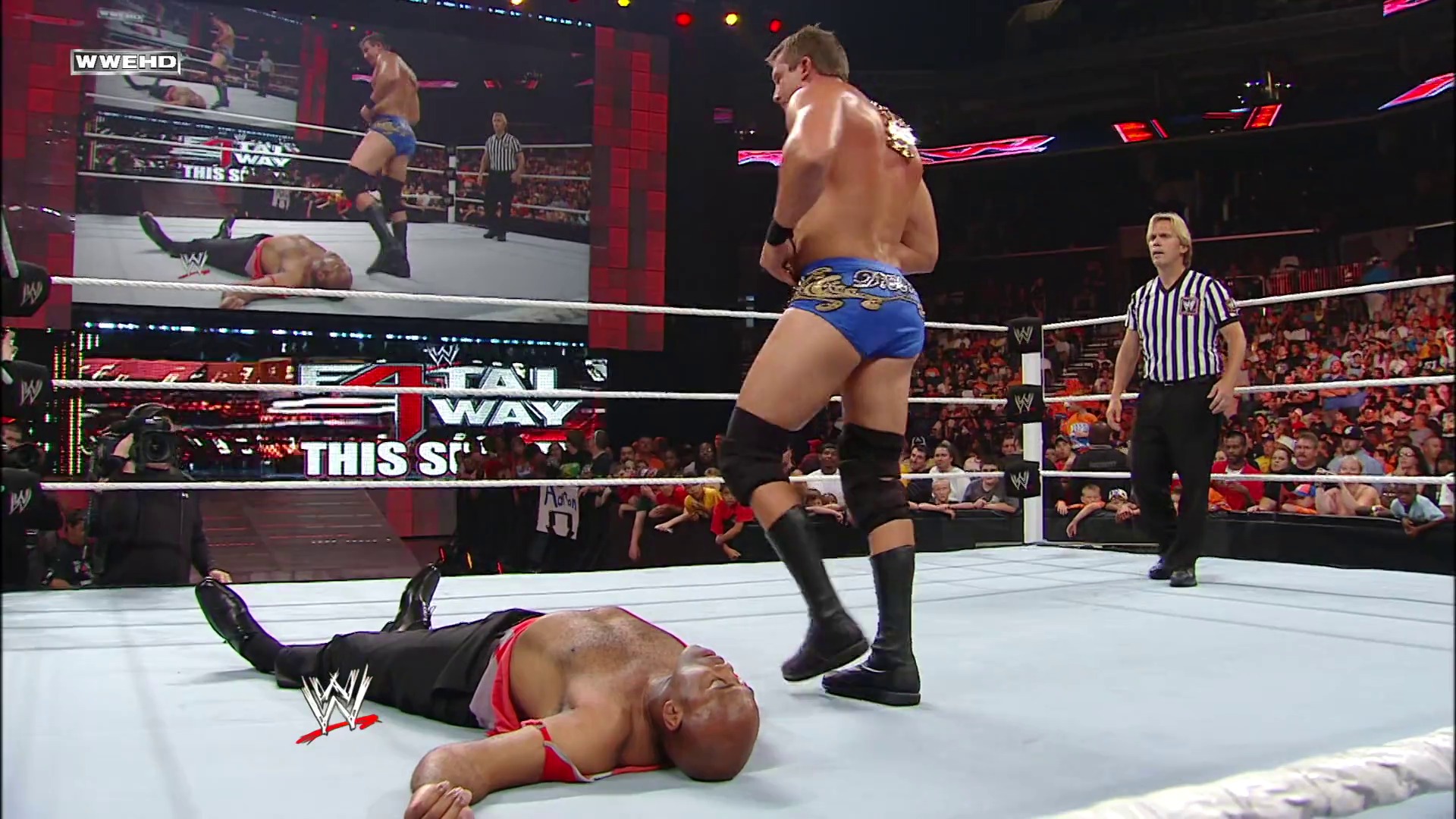 A frame from WWE Raw (June 14, 2010) at 1:05:08. A WWE Raw match in progress shows one wrestler lying on the white canvas while another stands nearby, with a referee in a striped shirt observing. The packed arena crowd is visible in the background beneath red-themed lighting and graphics.