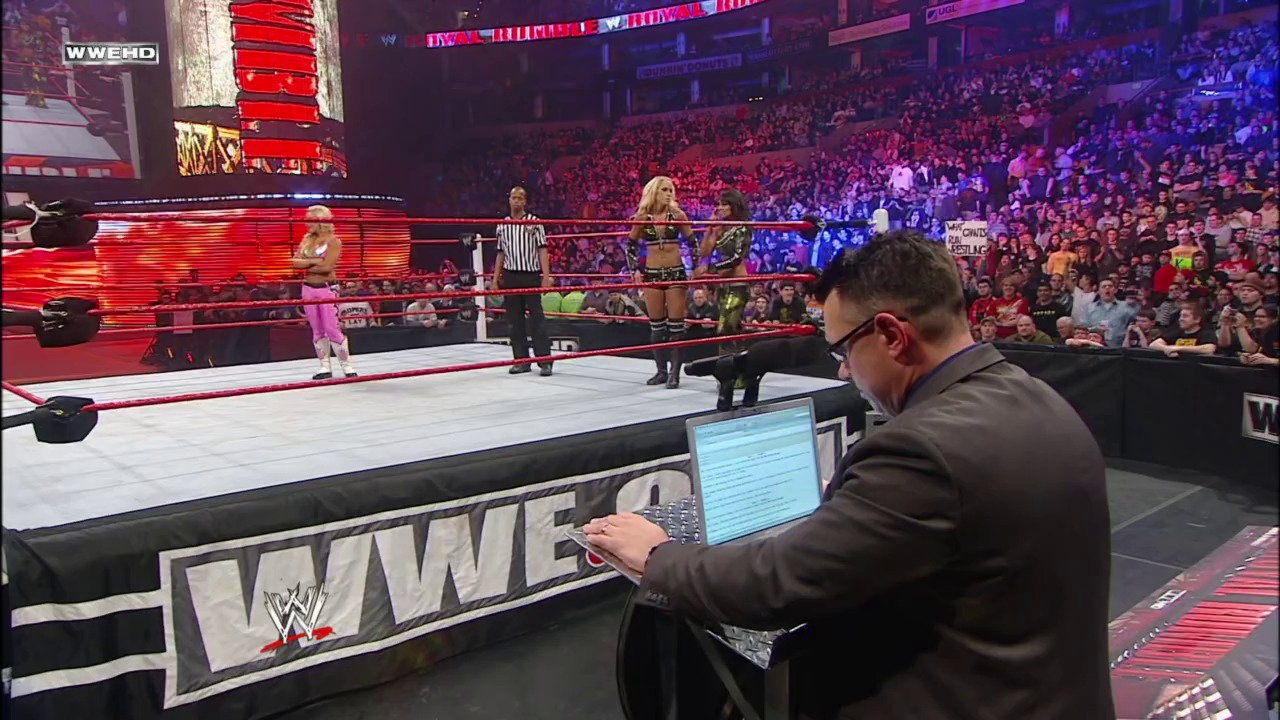 A frame from Royal Rumble (2011) at 1:10:19. A WWE official at ringside works on a laptop during a Royal Rumble match, while wrestlers and a referee are visible in the ring before a packed arena crowd under red and purple lighting.