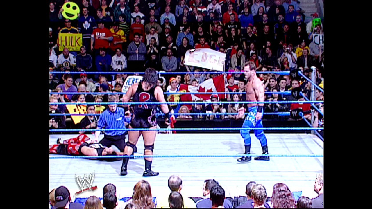 A frame from WWE SmackDown (February 27, 2003) at 37:46. A WWE SmackDown match in progress with a wrestler in the ring facing an opponent, while another competitor lies on the mat. The crowd fills the arena seats behind the blue-roped ring, with WWE branding and signage visible throughout the venue.