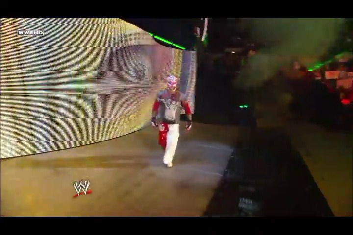 A frame from WWE Raw (July 25, 2011) at 1:12. A wrestler in red and white attire walks down the ramp toward the ring during a WWE Raw event, with a large video screen displaying graphics behind them and the WWE logo visible in the corner.