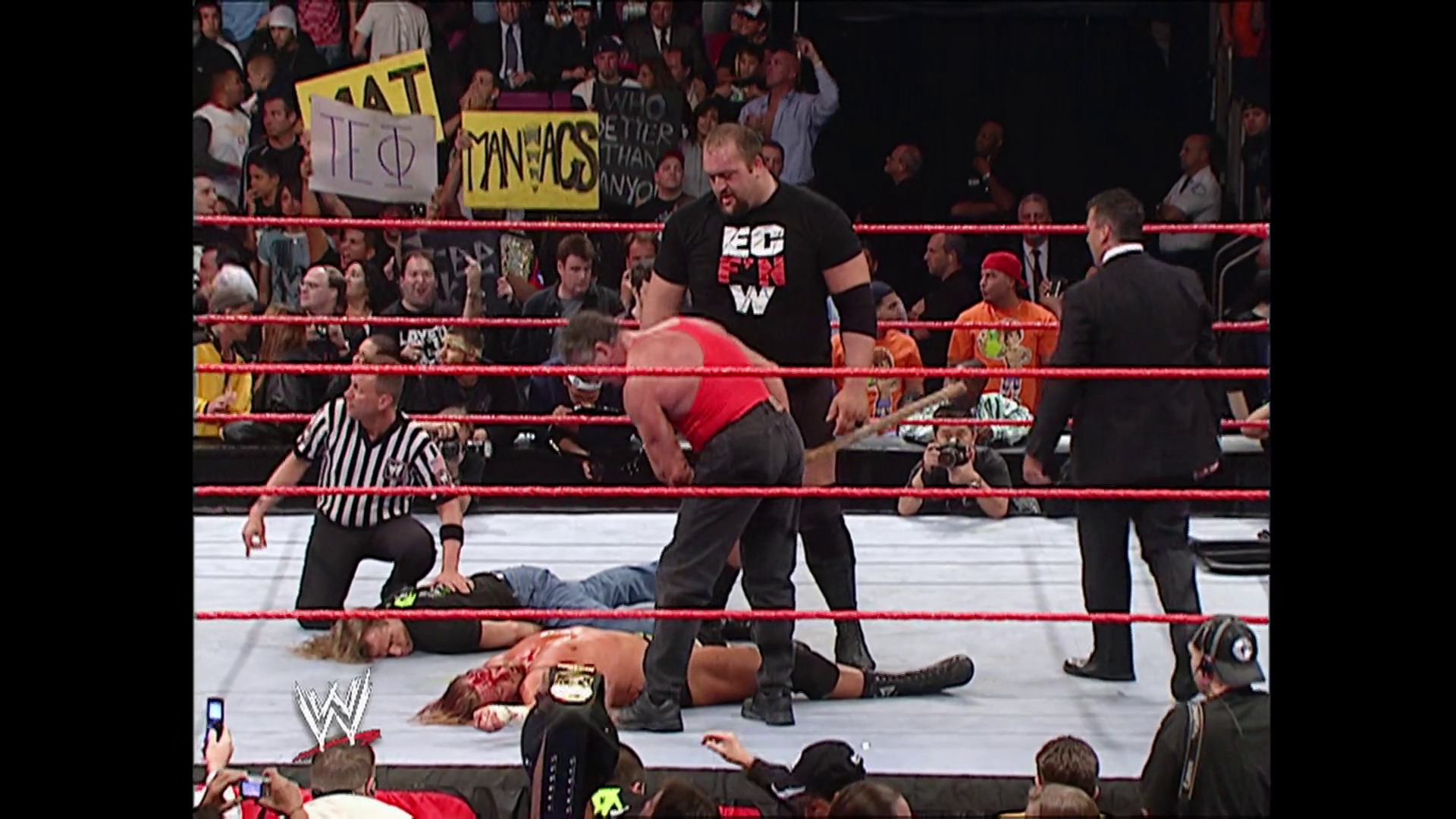 A frame from WWE Raw (September 11, 2006) at 1:30:54. A WWE Raw match in progress shows a wrestler in red and black standing in the ring while opponents lie on the mat. A referee in striped shirt oversees the action as the crowd watches from the stands behind the red ropes.