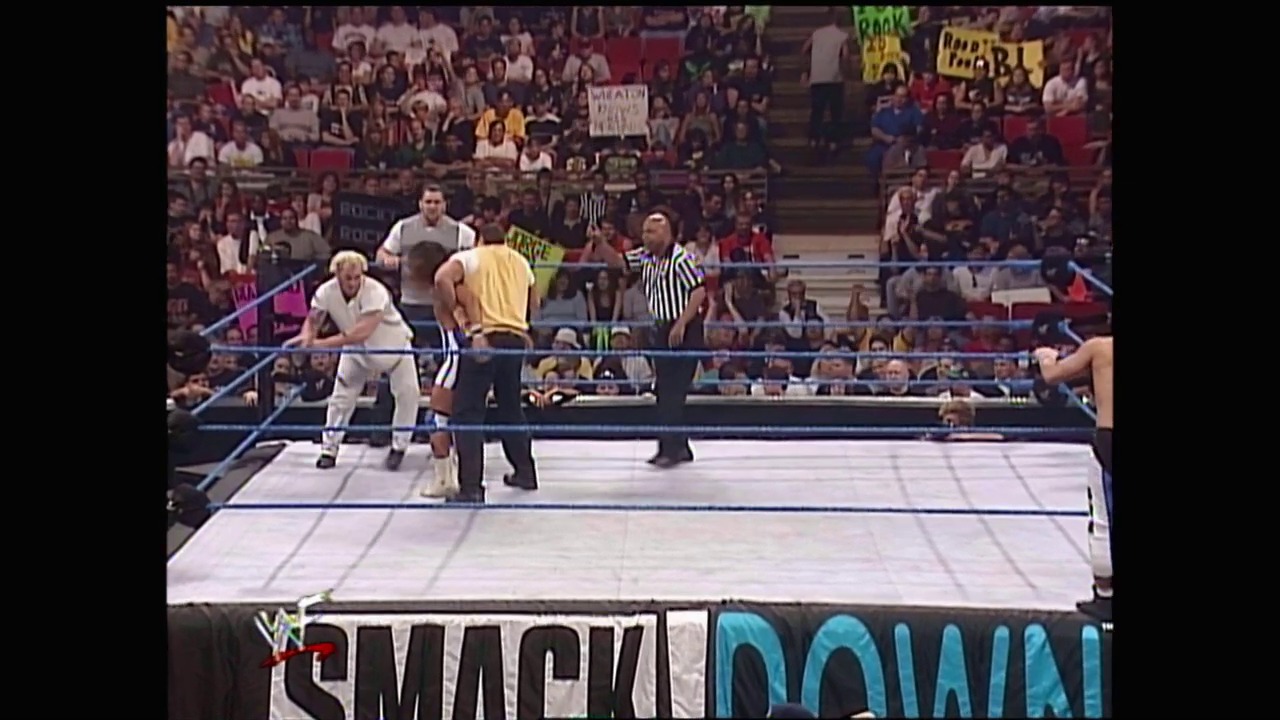 A frame from WWE SmackDown (January 6, 2000) at 1:07:18. # WWE SmackDown Match Scene

A wrestling match is underway in a packed arena with wrestlers in the ring—one in white attire and another in yellow—while a referee in stripes oversees the action. The crowd fills the bleachers in the background, and the "SmackDown" logo is visible at the bottom of the frame.