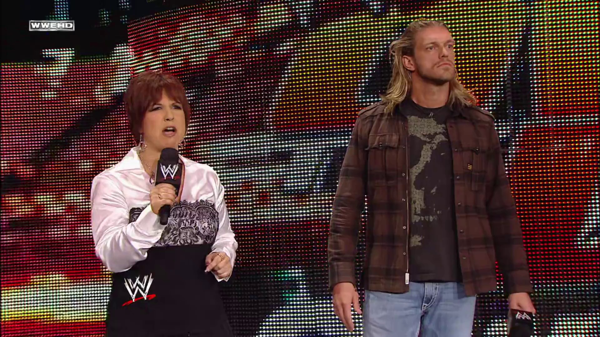 A frame from WWE Raw (May 10, 2010) at 11:19. A female WWE announcer in a white shirt holds a microphone while standing next to a blonde male wrestler in a brown leather jacket, with a colorful LED screen backdrop illuminating the arena during a WWE Raw broadcast.