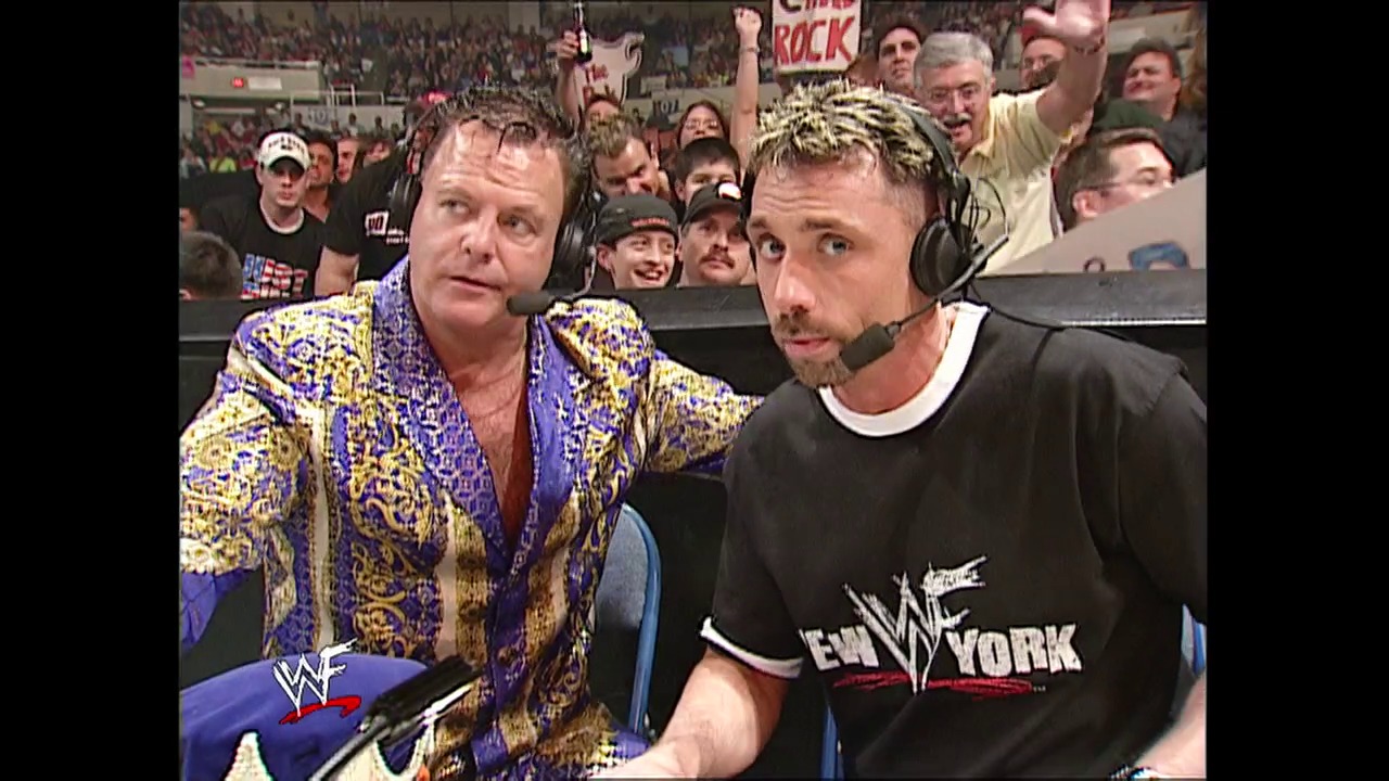 A frame from WWE Raw (January 31, 2002) at 27:11. Two commentators wearing headsets sit at ringside during a WWE Raw broadcast, with one in a distinctive blue and gold patterned jacket and the other in a black "New York" shirt. A large enthusiastic crowd fills the arena behind them.