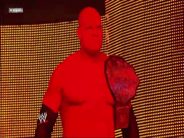 A frame from WWE SmackDown (October 29, 2010) at 1:22:02. A wrestler in red attire stands against a golden LED backdrop with the WWE logo visible. The figure is illuminated in red lighting against the warm-toned pixelated screen display.