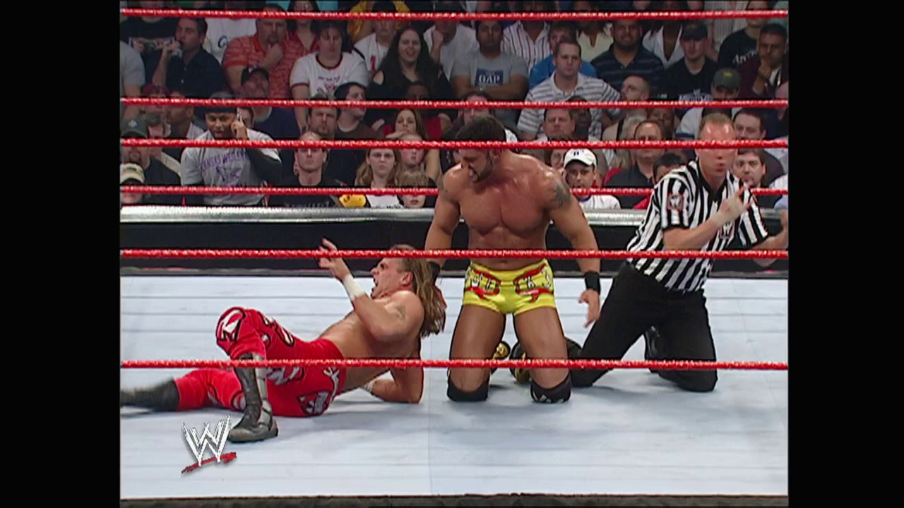 A frame from WWE Raw (March 28, 2005) at 1:05:06. A WWE wrestler in red attire kneels on the mat while an opponent stands over him in the ring. A referee in black and white stripes watches the action as the crowd fills the stands behind the red ropes.
