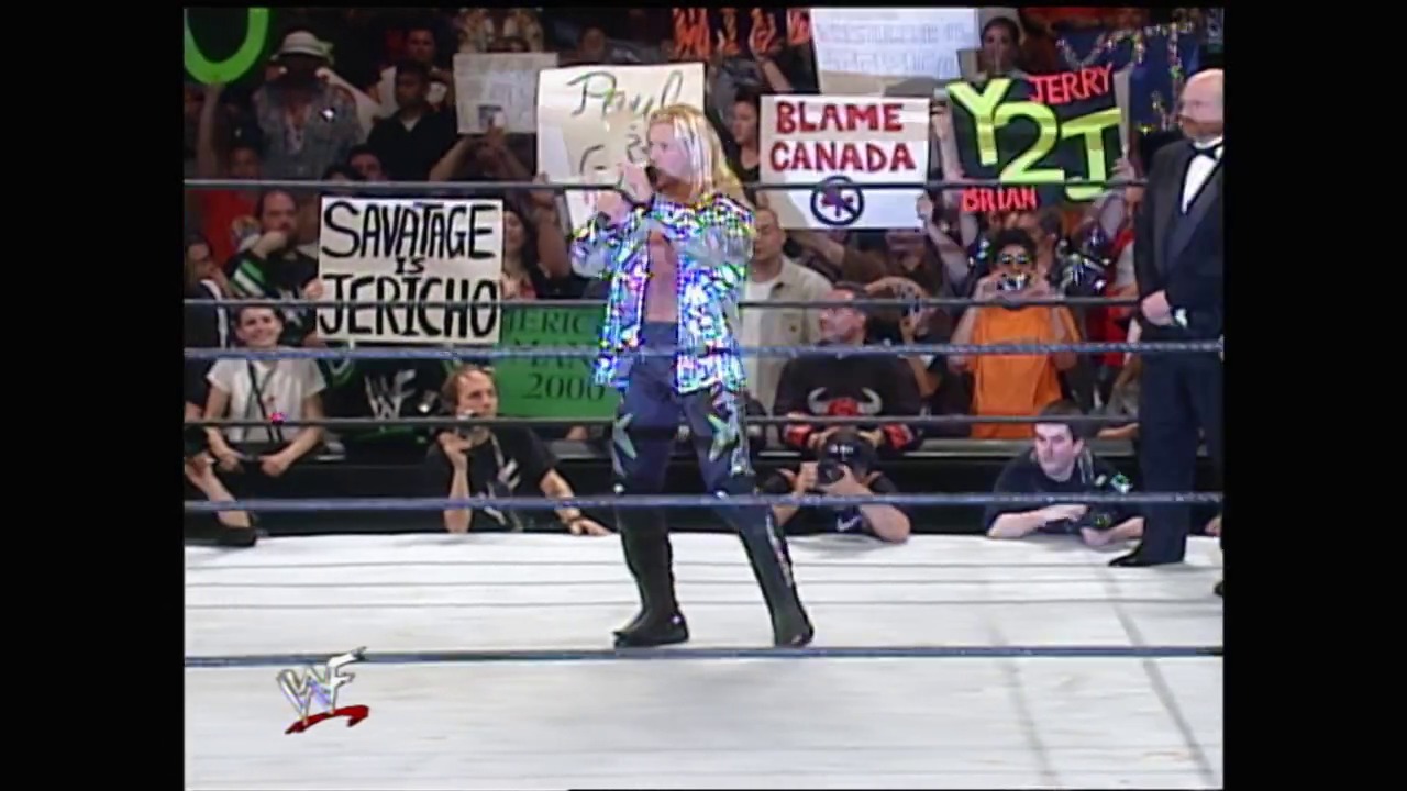 A frame from WrestleMania 2000 (2000) at 1:52:56. A blonde female wrestler in a colorful blue and white outfit stands in the WWF wrestling ring, with enthusiastic fans holding signs behind her including "BLAME CANADA" and "SAVAGE JERICHO." The crowd and ringside personnel are visible in the background.