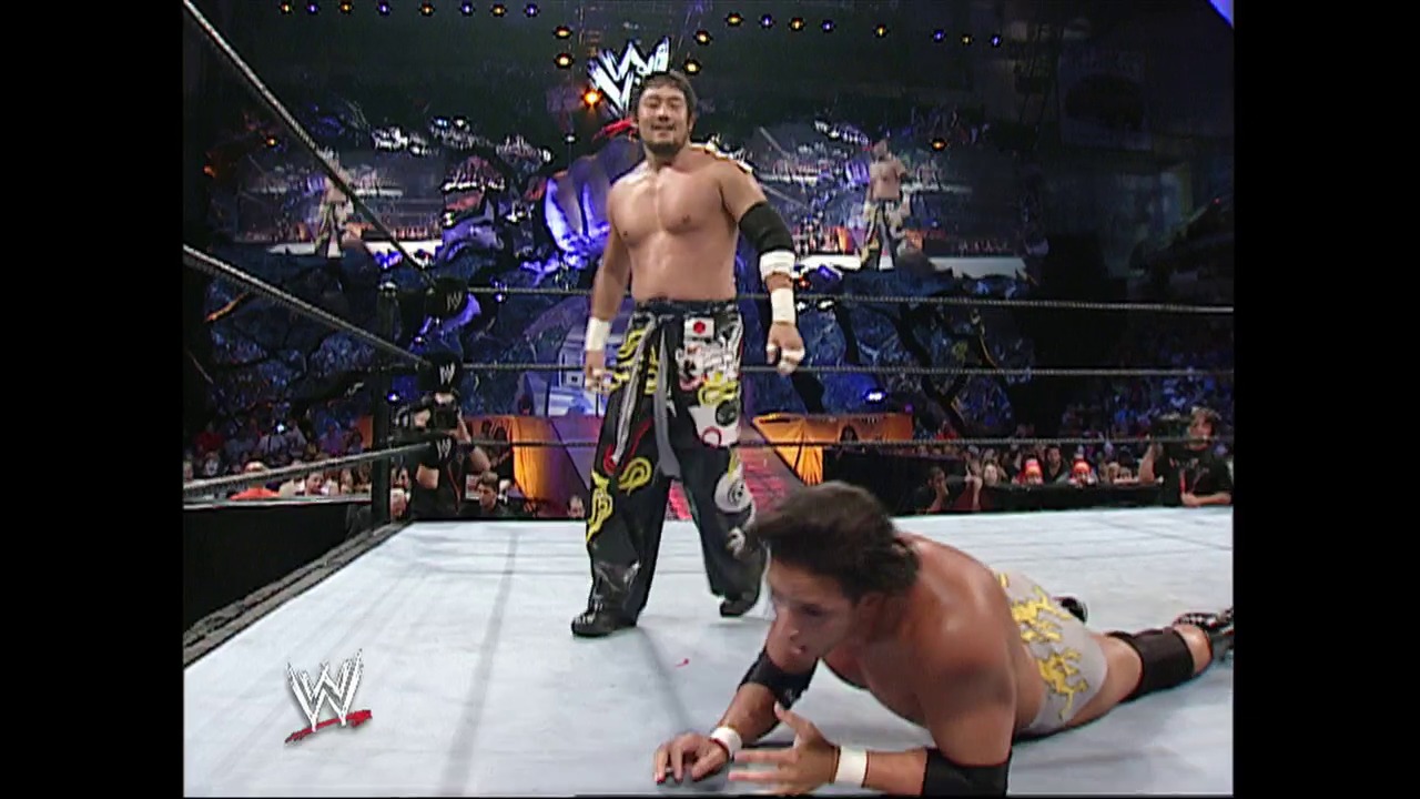 A frame from WWE Raw (July 18, 2002) at 1:01:27. A WWE wrestler stands triumphantly in the ring wearing distinctive black and gold patterned pants, while an opponent lies on the white mat below. The packed arena crowd is visible in the background with the illuminated WWE logo overhead.