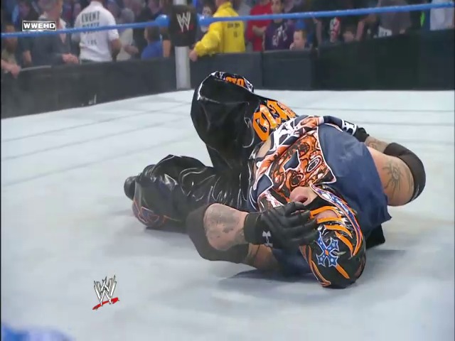A frame from WWE SmackDown (January 22, 2010) at 1:22:36. A wrestler lies on the mat in a striking orange and blue outfit during a WWE SmackDown match, with the crowd visible in the background and the WWE logo displayed at ringside.