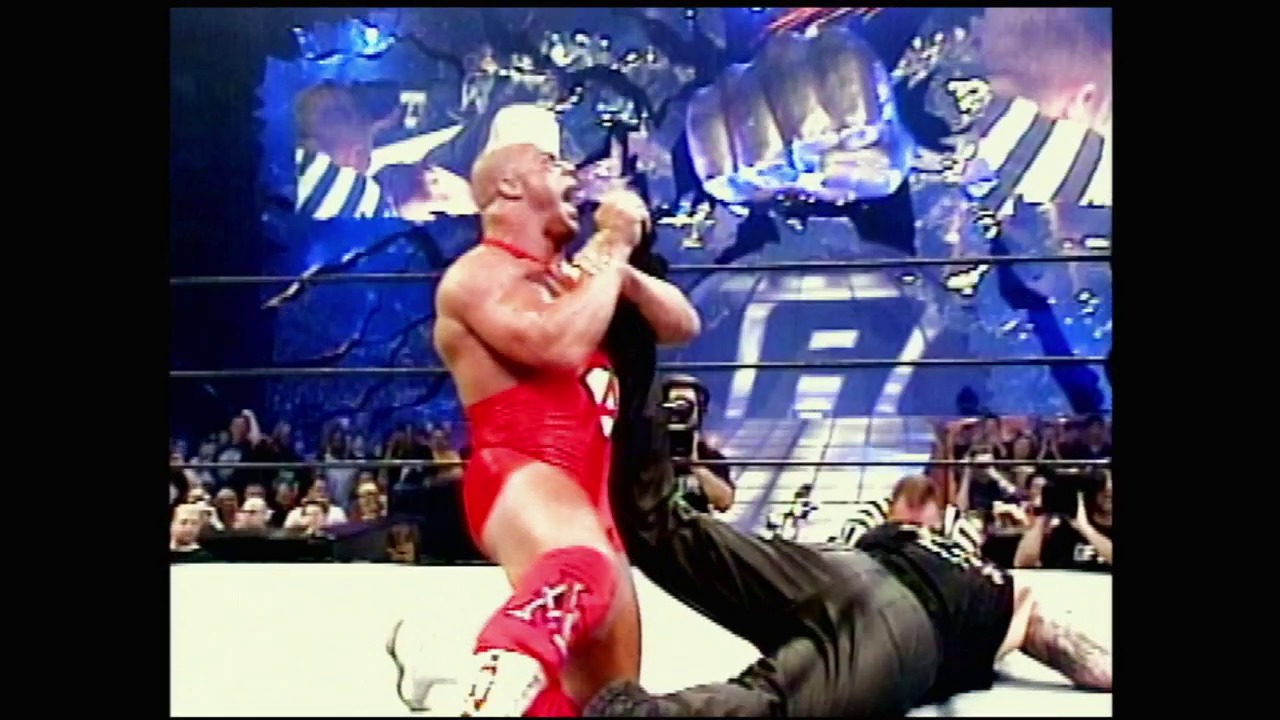 A frame from WWE Vengeance (2002) at 0:48. A muscular wrestler in red attire and white headband performs in a WWE ring, with the crowd visible at ringside and blue graphics displayed on the arena's large screens behind the action.