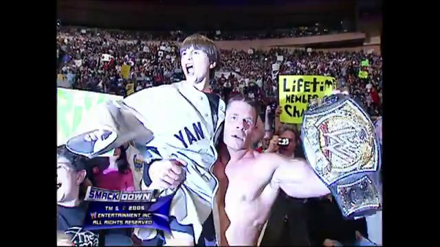A frame from WWE SmackDown (April 22, 2005) at 1:27:30. A shirtless wrestler holds a championship belt while posing with a cardboard cutout in a packed arena during WWE SmackDown. The crowd fills the background as WWE graphics and signage are visible around the ring.