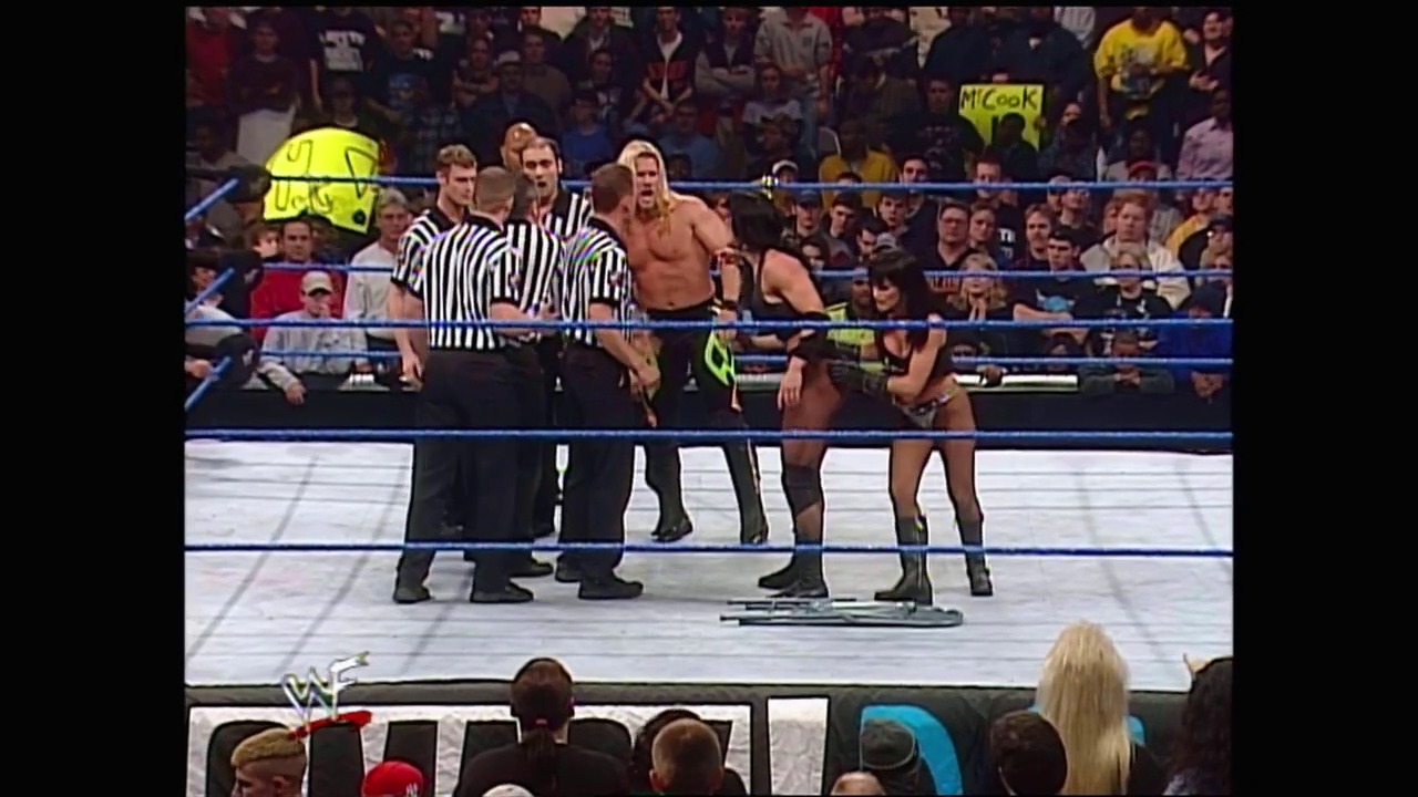 A frame from WWE SmackDown (December 30, 1999) at 1:03:57. # WWE SmackDown December 30, 1999

A wrestling match in progress inside a blue-roped ring with referees in striped shirts overseeing the action. Wrestlers in the ring face a packed crowd, with visible WWE graphics and audience members in the foreground wearing various colored attire.