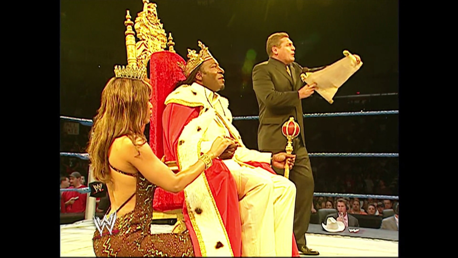 A frame from WWE SmackDown (July 14, 2006) at 4:09. # WWE SmackDown Ring Scene

A WWE wrestler in elaborate red and gold royal attire sits in the ring while a female performer stands beside him and an official in a brown suit holds documents. Ornate throne-like set pieces and crowd spectators are visible in the background.
