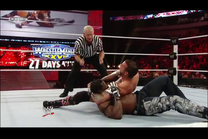 A frame from WWE Raw (March 7, 2011) at 1:07:01. A WWE wrestler executes a submission move on an opponent in the ring while a referee in striped shirt oversees the match. The arena is filled with spectators, and WrestleMania promotional graphics are visible on the displays.