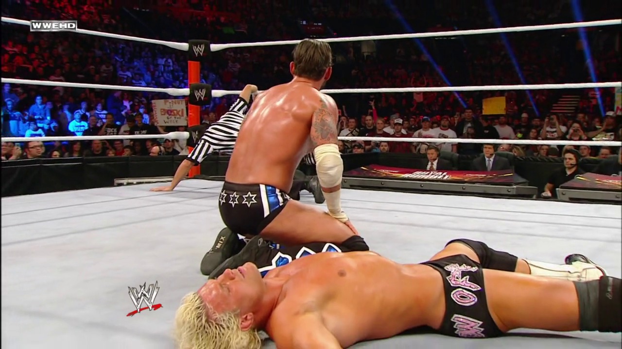 A frame from Royal Rumble (2012) at 1:32:50. A WWE wrestler in star-patterned trunks dominates an opponent on the canvas during a Royal Rumble match, with the referee and enthusiastic crowd visible in the background of the illuminated ring.