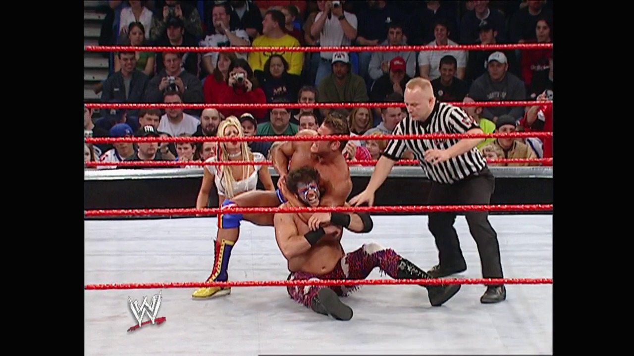 A frame from WWE Raw (February 2, 2004) at 27:40. A WWE wrestler in maroon and black attire is being thrown or falling in the ring while a referee in black and white stripes watches nearby. The red-roped ring is surrounded by a crowd of spectators, with the WWE logo visible in the lower left corner.