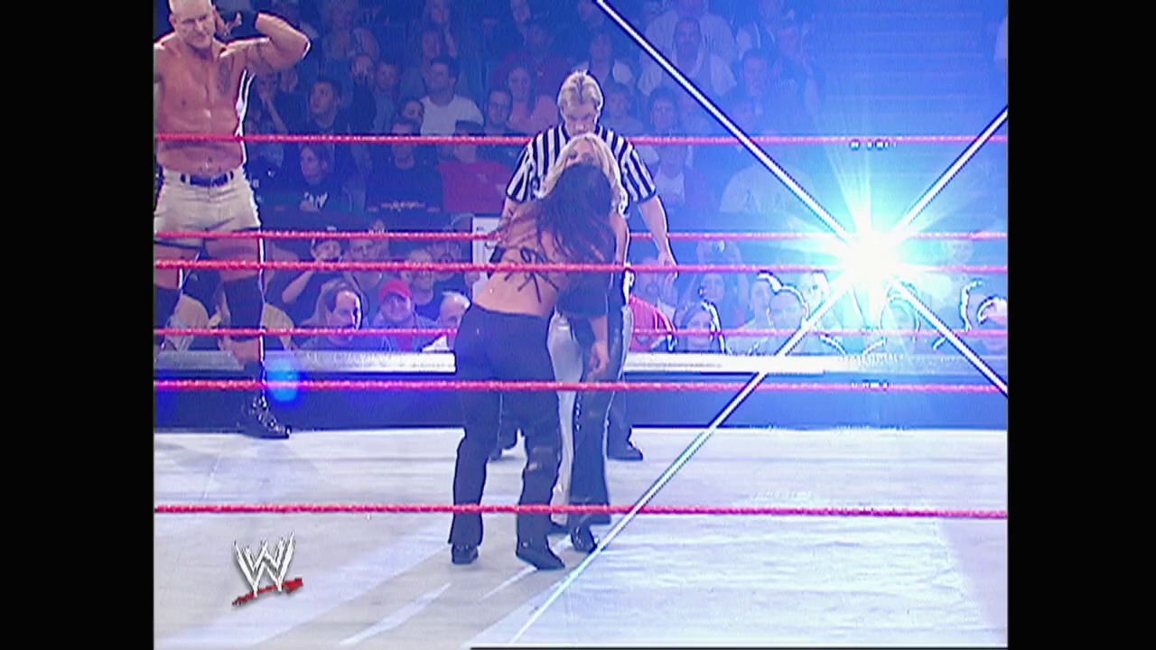 A frame from WWE Raw (November 3, 2003) at 1:04:27. A WWE Raw match in progress featuring a referee in a striped shirt overseeing two wrestlers in the red-roped ring, with an enthusiastic crowd visible in the background and the WWE logo displayed in the lower left corner.