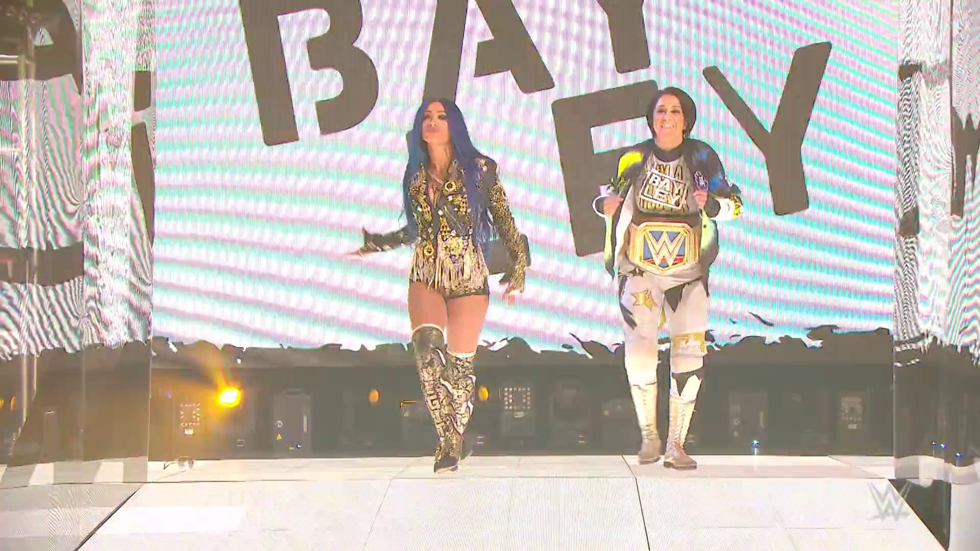 A frame from WWE SmackDown (June 5, 2020) at 1:13:17. Two female wrestlers make their entrance on a WWE SmackDown stage, with one wearing a gold-sequined jacket and the other displaying a championship belt. Large video screens behind them display WWE branding as they walk down the illuminated ramp.