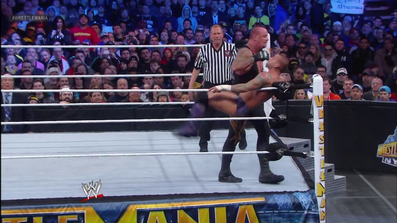 A frame from WrestleMania 29 (2013) at 2:09:02. A wrestler executes a move on his opponent in the WWE ring during WrestleMania 29, with a referee in striped shirt overseeing the match. A massive crowd fills the arena while the WrestleMania logo is visible on the ring apron.