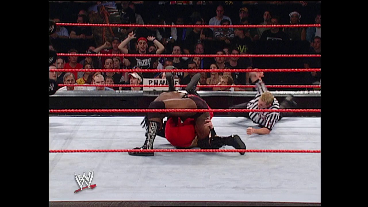 A frame from WWE Raw (July 7, 2003) at 14:15. A WWE wrestler in red lies on the mat while a referee in a striped shirt stands nearby. The red-roped wrestling ring is surrounded by an enthusiastic crowd, with the WWE logo visible in the lower left corner.