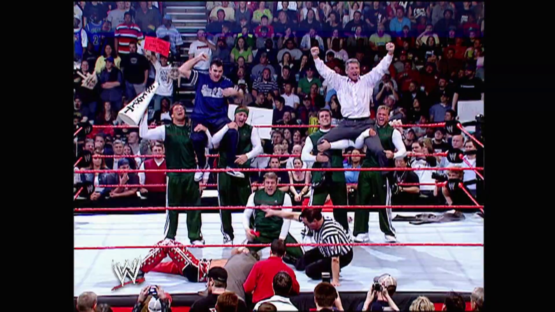 A frame from WWE Raw (March 6, 2006) at 49:53. A WWE Raw match in progress shows a referee attending to a fallen wrestler in the red ring, while another wrestler celebrates with arms raised at the ropes. Spectators fill the arena stands surrounding the action.