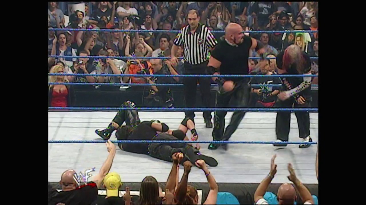 A frame from WWE SmackDown (July 20, 2000) at 51:11. A wrestler lies on the mat during a WWE SmackDown match, with a referee in striped shirt overseeing the action. The crowd cheers enthusiastically from the stands surrounding the blue-roped wrestling ring.