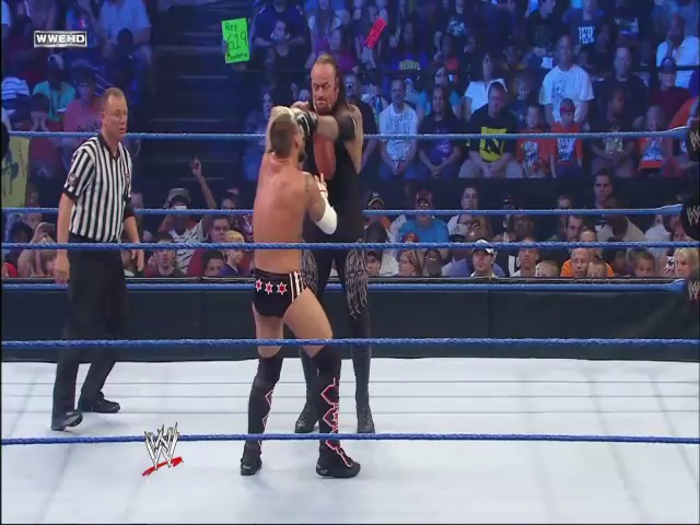 A frame from WWE SmackDown (September 10, 2010) at 1:08:11. Two wrestlers are engaged in combat in a WWE ring, with one delivering a punch to his opponent while a referee in a striped shirt watches. The crowd fills the arena in the background, and the blue WWE branding is visible around the ring.