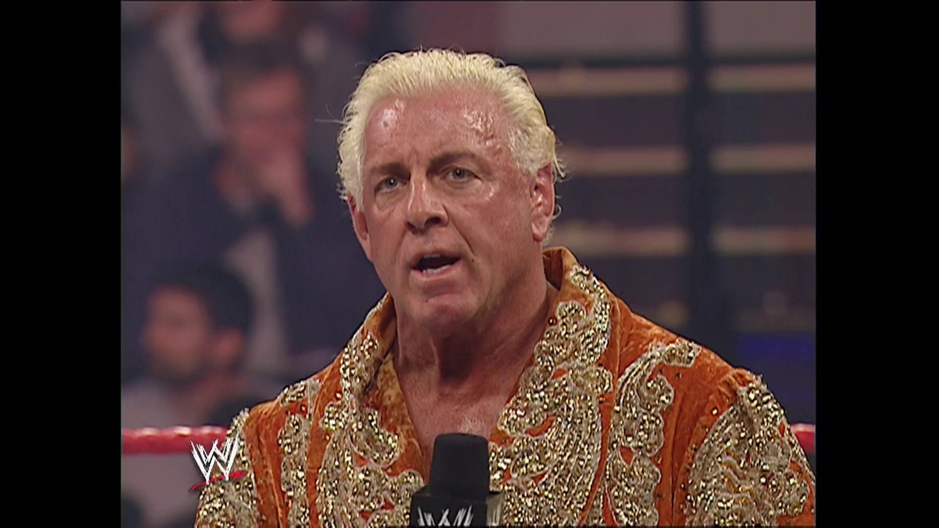 A frame from WWE Raw (November 27, 2006) at 3:08. A professional wrestler with white/blonde hair wears an ornate orange and gold robe while speaking into a microphone in a WWE ring. The WWE logo is visible in the lower left corner, with blurred crowd members visible in the background.