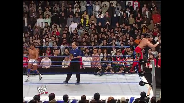 A frame from WWE SmackDown (February 11, 2005) at 25:28. A WWE wrestler in red trunks performs an aerial move from the ring ropes while opponents and a referee watch below, with a large crowd visible in the background of the packed arena.