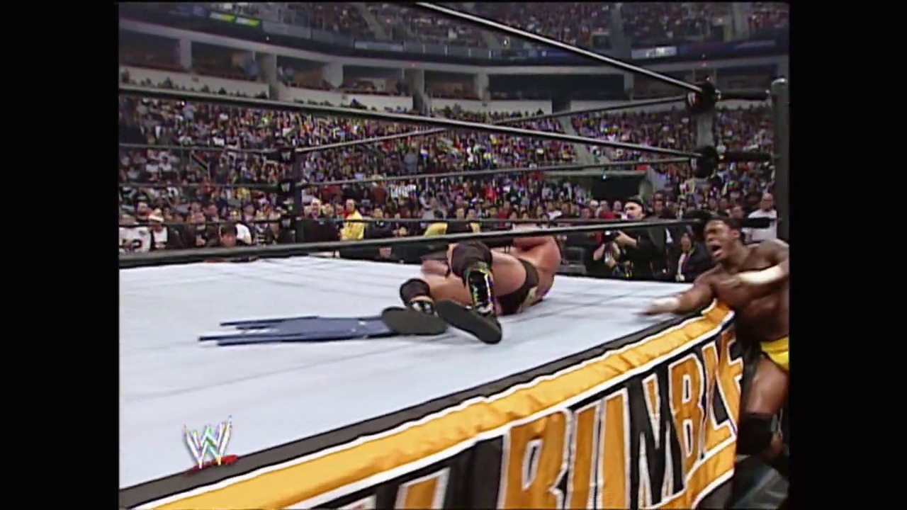 A frame from WWE Royal Rumble (2005) at 1:14:25. A wrestler lies on the mat in a WWE ring during Royal Rumble 2005, with another wrestler visible at ringside. The packed stadium crowd fills the background while the yellow ring apron displays the event branding.