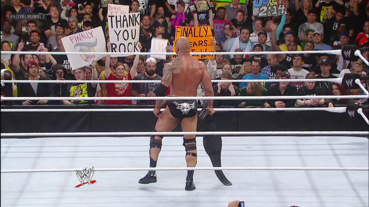 A frame from Royal Rumble (2013) at 2:45:43. A shirtless wrestler stands in a WWE ring facing an enthusiastic crowd holding signs, including one reading "THANK YOU ROCK." The wrestler displays a bull tattoo on his chest while fans cheer behind the white ropes.